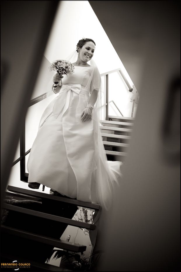 The bride, bouquet in hand, comes down the wooden stairs, smiling, to leave for the wedding ceremony in Seia, Portugal.
