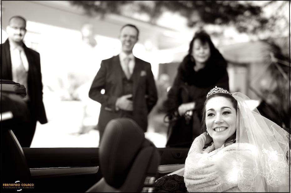 With the groom and a couple of guests in the background and out of focus, the bride smiles while still sitting in the car when she arrives at Quinta do Joinal in Caparica for the wedding ceremony.