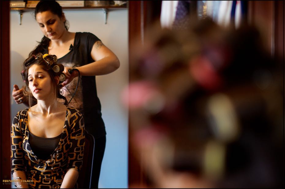 Seated, the bride having her hair done, with the hairdresser behind her, seen in a mirror in a composition by the wedding photographer in Cascais.