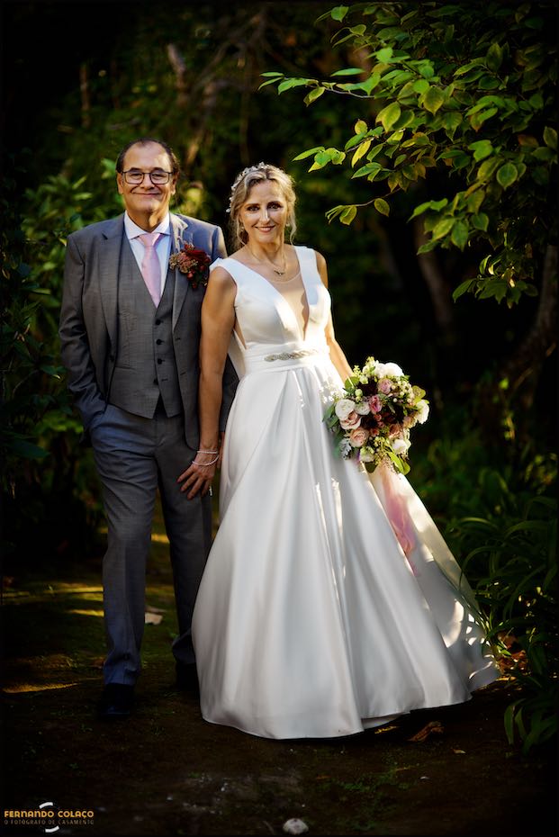 Inside the trees of the garden of Quinta do Pé da Serra in Sintra, the bride, standing and bouquet in hand, leans against the groom, in a composition by the wedding photographer in Sintra.