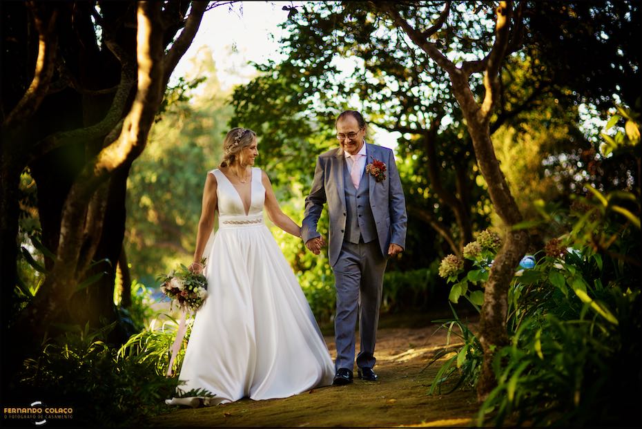 Among the large trees in the garden of Quinta do Pé da Serra, the bride and groom walk hand in hand, smiling and looking at each other, as seen by the wedding photographer in Sintra.